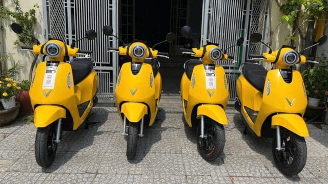 Four yellow scooters are parked in front of a Hoi An building with a gated entrance and some plants nearby.
