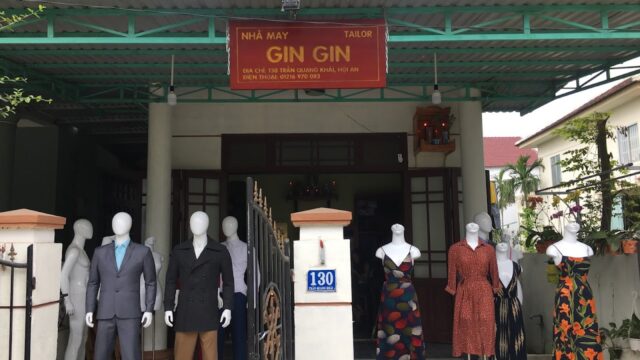 Mannequins in formal and casual outfits stand at a Hoi An tailor shop entrance, beneath a bold red sign.