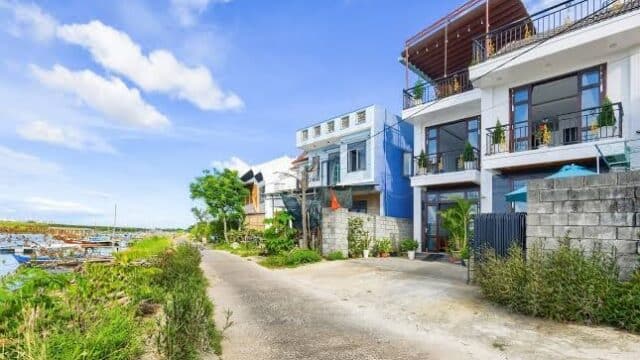 A quiet street in Hoi An with modern houses and boats by the water under a bright blue sky with scattered clouds.
