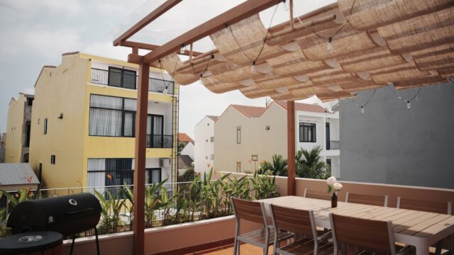 Outdoor patio with wooden table, chairs, and pergola evokes a Hoi An vibe amid modern buildings and lush plants in the background.
