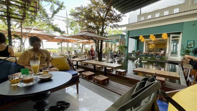 Outdoor café in Hoi An with benches, drinks, pond, umbrellas, and lush greenery. Bright and relaxing atmosphere for all.