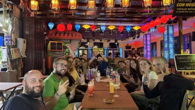 A group of friends enjoying drinks at a lively, colorful outdoor restaurant at night in vibrant Hoi An.