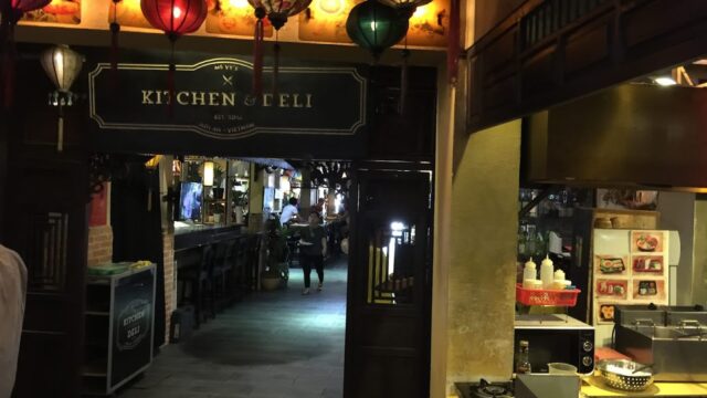 Dimly lit deli entrance in Hoi An with hanging lanterns, a long bar, and a kitchen counter displaying food photos.