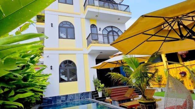 A modern three-story house in Hoi An with balconies, a pool, tropical plants, lounge chairs, and a large yellow patio umbrella.
