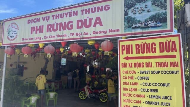 A coconut boat tour shop in Hoi An with a yellow sign, red lanterns, and a menu offering coffee, lemon juice, and ice cream.
