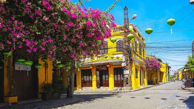 A sunny Hoi An street with bright yellow buildings, pink bougainvillea vines, and green lanterns hanging above.