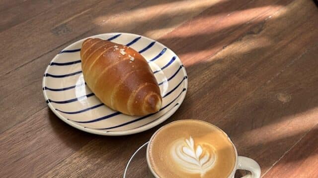 A cup of latte with latte art and a croissant on a striped plate, set on a sunny wooden table in charming Hoi An.