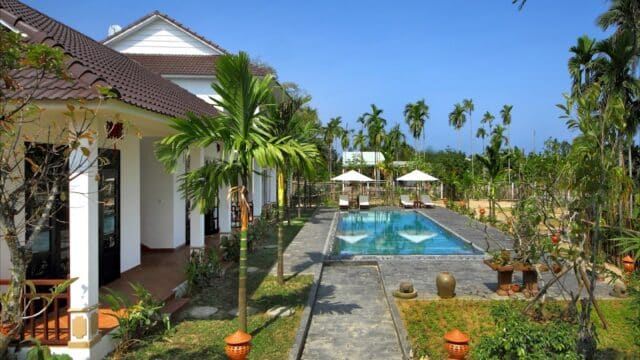 A white Hoi An villa with a pool, palm trees, and sun loungers under umbrellas in a lush garden on a sunny day.