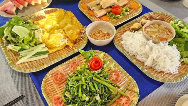 A table set with Hoi An Vietnamese dishes, fresh vegetables, rice noodles, dipping sauce, and sliced watermelon on woven trays.