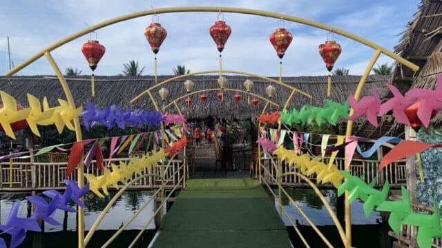 A decorated bridge in Hoi An with colorful pinwheels, lanterns, and streamers leads to thatched huts over water.