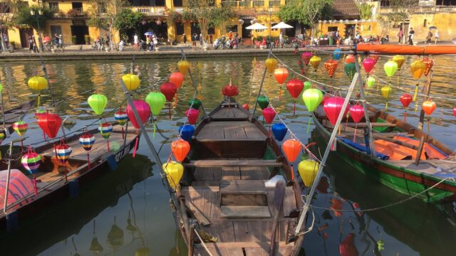Wooden boats with colorful lanterns float on Hoi An’s river, with yellow buildings and people in the background.