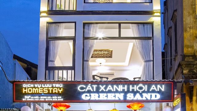 A brightly lit, modern three-story homestay building with large windows and a Green Sand sign welcomes you in Hoi An.