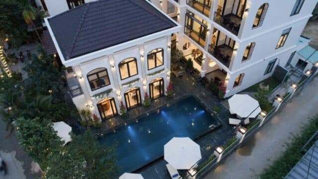 Aerial view of a modern white hotel with balconies and a lit outdoor pool surrounded by umbrellas in Hoi An.