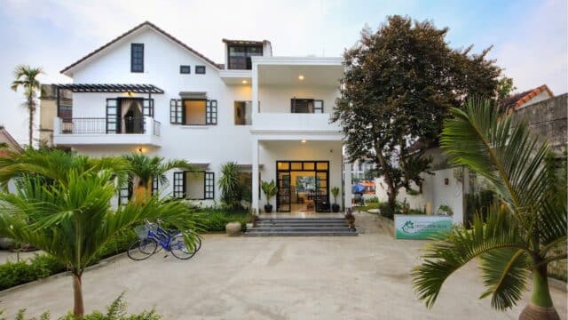 White two-story house in Hoi An with balconies, large windows, a tree, potted plants, and bicycles parked in the front driveway.