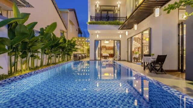 Modern Hoi An house with large blue-tiled pool, outdoor seating, lush plants, and warm lights glowing at dusk.
