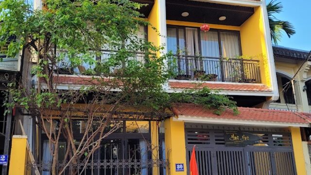 A three-story yellow building with balconies in Hoi An, featuring a gated entrance and a tree in front on a sunny day.