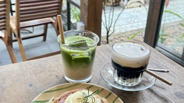 A cinnamon roll with lemon, a matcha drink, and coffee sit on a wooden table in Hoi An by a window with a garden view.