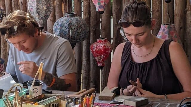 Two people sit at a colorful craft table making art, inspired by Hoi An, with decorated lanterns and wooden sticks in the background.
