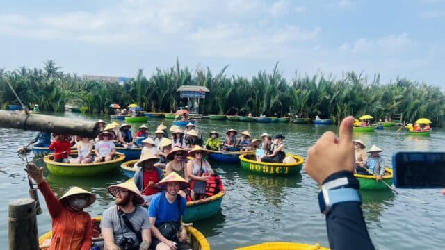 Groups of people wearing hats sit in round basket boats on a river in Hoi An, surrounded by palm trees under a blue sky.