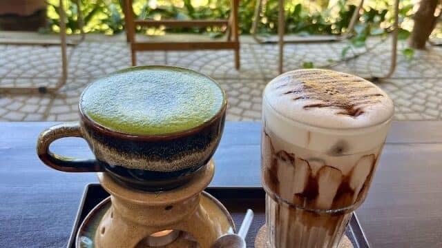 Enjoy a Matcha latte and iced coffee with cream, served on a tray in Hoi An, surrounded by chairs and lush greenery.
