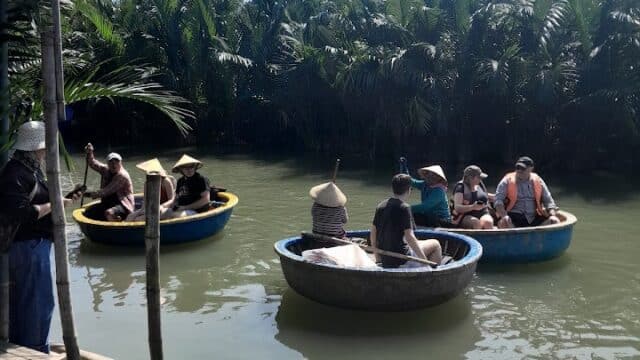 People in round basket boats paddle on a river near Hoi An, surrounded by lush greenery under a clear sky.