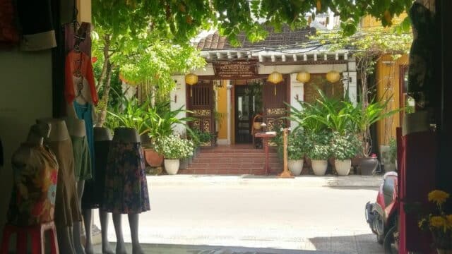 A street view in Hoi An of a boutique with mannequins outside, facing a traditional building adorned with potted plants and lanterns.
