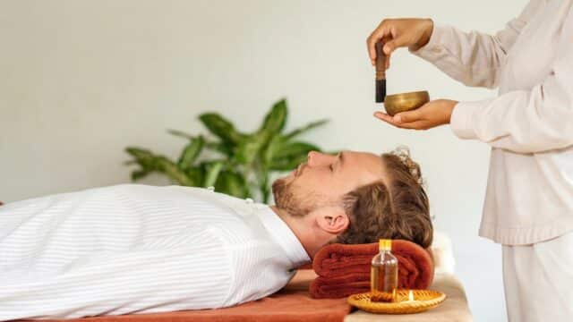 A person lies on a massage table in Hoi An as another holds a singing bowl and mallet by their head, suggesting a sound healing session.