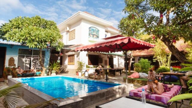People relax by a small backyard pool in Hoi An, surrounded by trees and buildings on a sunny day.
