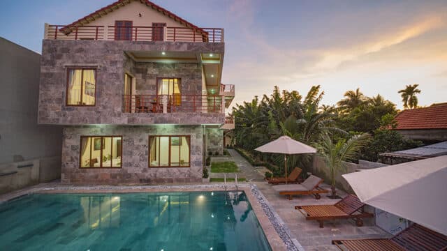 Modern two-story house with stone exterior, pool, and lush greenery evokes a Hoi An retreat at sunset.
