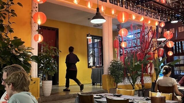 Outdoor Hoi An restaurant with red lanterns, yellow walls, and diners seated at tables under warm string lights at night.