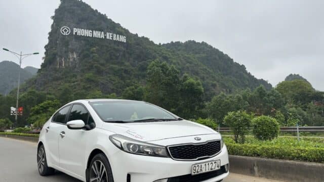 A white Kia parked on a road near green hills, with a Phong Nha-Ke Bang sign and Hoi An vibes in the scenic background.