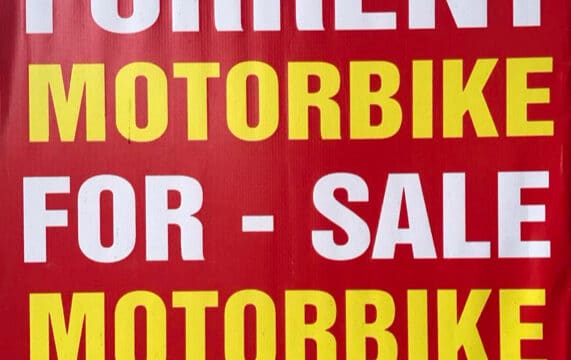 Red and yellow sign in Hoi An offering motorbikes for rent and sale, plus laundry service, with contact info and address at bottom.