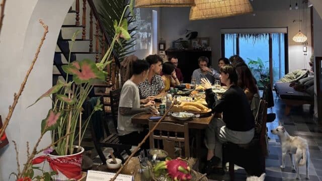 A group shares a meal at a cozy home in Hoi An, with two cats nearby and warm hanging lights above the dining table.