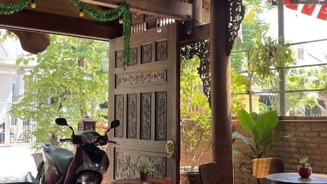 A cozy Hoi An cafe with wooden furniture, open door, sunlight, and a motorbike parked outside near lush plants.
