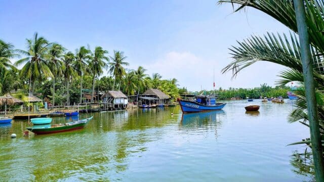 Colorful boats float on a calm river in Hoi An, lined with palm trees and thatched huts under a clear blue sky.
