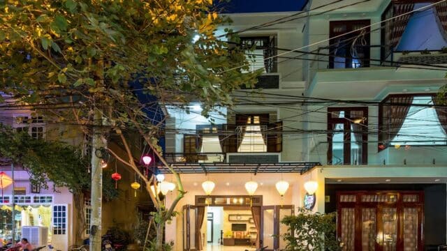 Three-story Hoi An building at night with lanterns, balconies, parked scooters, and a large tree in front.