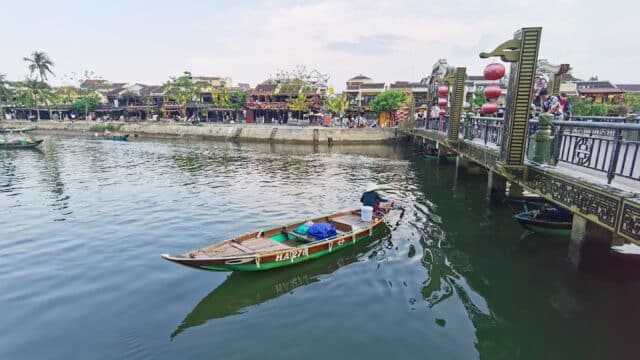 A person rows a small wooden boat on a river near Hoi An’s decorated bridge and charming riverside buildings.