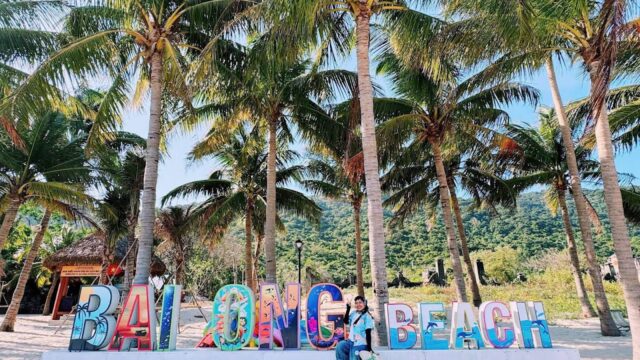 Colorful BALONG BEACH sign near Hoi An, with a person relaxing under palm trees on a sunny, tropical beach day.