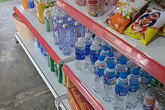 Store shelves in Hoi An stocked with bottled water, instant noodles, snacks, canned drinks, and various packaged foods.