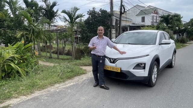 A man stands beside a white car on a quiet street in Hoi An, with houses, plants, and power lines lining the background.