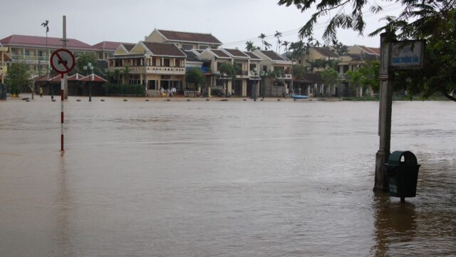 Flooded Hoi An street with water reaching building entrances, partially submerging a sign and trash bin.