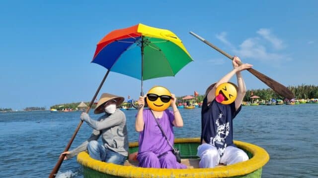 Three people sit in a round boat on Hoi An waters; one holds a rainbow umbrella. Two faces have emoji overlays. Clear blue sky above.