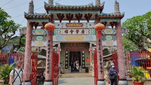 People walk near the entrance of a traditional Hoi An Chinese temple adorned with red lanterns and ornate decorations.