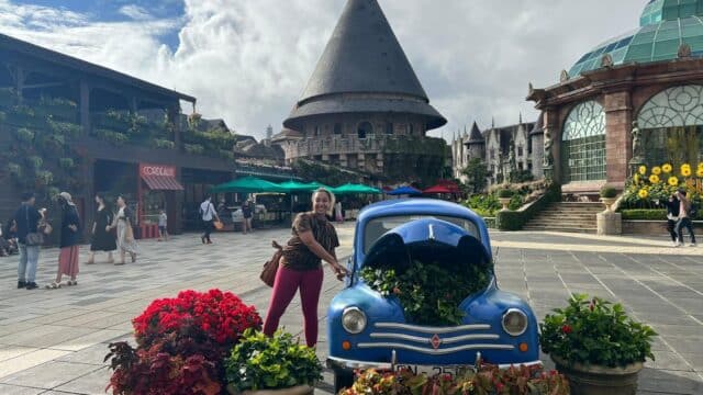 A woman poses by a blue car planter in Hoi An square, surrounded by flowers, people, and unique buildings under a partly cloudy sky.