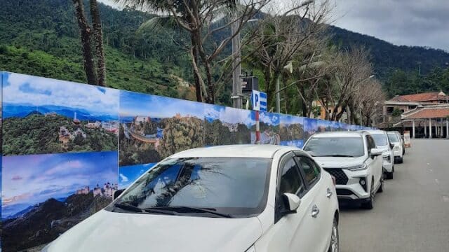 Four white cars are parked along a palm-lined road in Hoi An, with scenic mountains and a cloudy sky in the background.