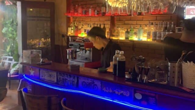 A bartender works behind a colorful, neon-lit Hoi An bar with empty stools and bottles displayed on shelves.