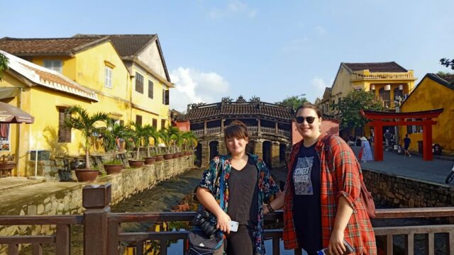 Two people stand on a bridge in Hoi An, with a historic Japanese bridge and yellow buildings beneath a clear blue sky.