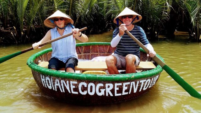 Two people in a round bamboo boat wearing conical hats paddle on a Hoi An river, framed by palm trees in the background.
