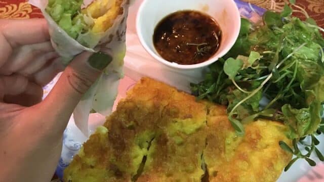 Hand holding a piece of Vietnamese bánh xèo with lettuce, beside dipping sauce and greens on a plate in Hoi An.