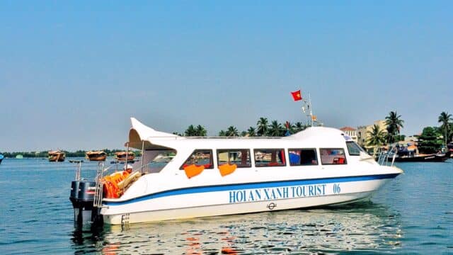 White passenger boat with HOI AN XANH TOURIST on its side floats on calm Hoi An waters, palm trees and buildings in the background.